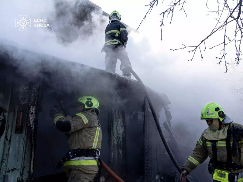 Три пожежники ДСНС Києва ліквідовують загоряння будівлі. Один рятувальник перебуває на даху з пожежним рукавом, двоє інших працюють біля входу в сильно задимлене приміщення.