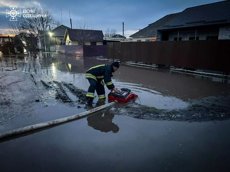 Рятувальники ДСНС Одещини відкачують воду із затопленого дитячого майданчика за допомогою мотопомпи. На фото видно пожежні автомобілі, рятувальників та кригу на підтопленій території. В лівому верхньому куті логотип ДСНС Одещини.