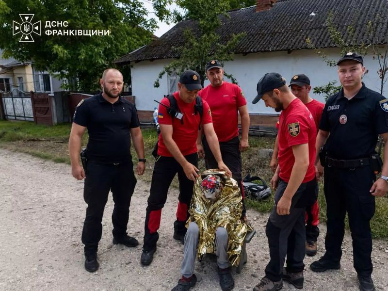 Рятувальники ДСНС та поліцейські стоять поруч зі знайденою 72-річною жінкою в золотистій термоковдрі після завершення пошуків на Прикарпатті.
