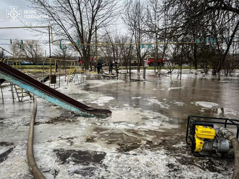 Рятувальники ДСНС Одещини відкачують воду із затопленого дитячого майданчика за допомогою мотопомпи. На фото видно пожежні автомобілі, рятувальників та кригу на підтопленій території. В лівому верхньому куті логотип ДСНС Одещини.